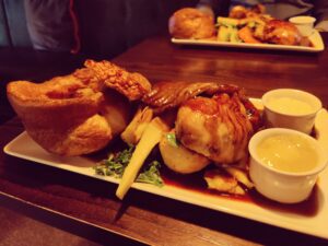 A plate of food featuring roasted vegetables, Yorkshire pudding, and a generous portion of meat drizzled with gravy, accompanied by two small pots of sauce. Another similarly arranged plate is visible in the background on a wooden table.